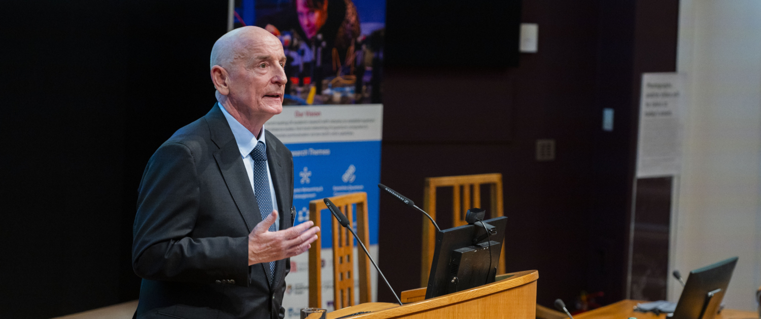 Image showing the Hub Director, Professor Gerald Buller (a white, middle-aged man) addressing the audience in a lecture theatre at the Hub launch event