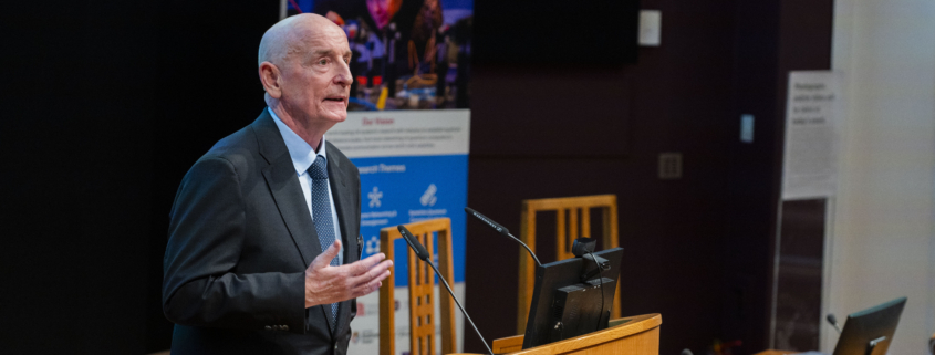 Image showing the Hub Director, Professor Gerald Buller (a white, middle-aged man) addressing the audience in a lecture theatre at the Hub launch event