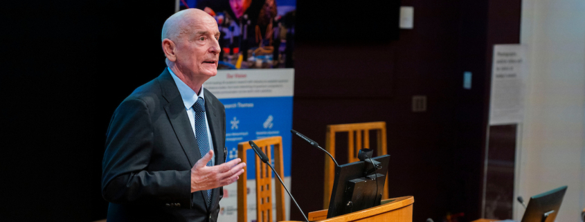 Image showing the Hub Director, Professor Gerald Buller (a white, middle-aged man) addressing the audience in a lecture theatre at the Hub launch event